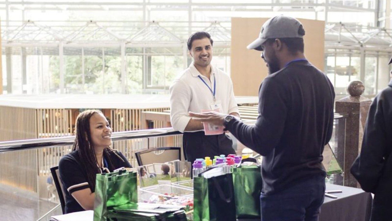 One person sits at a table with green bags and talks to a standing person
