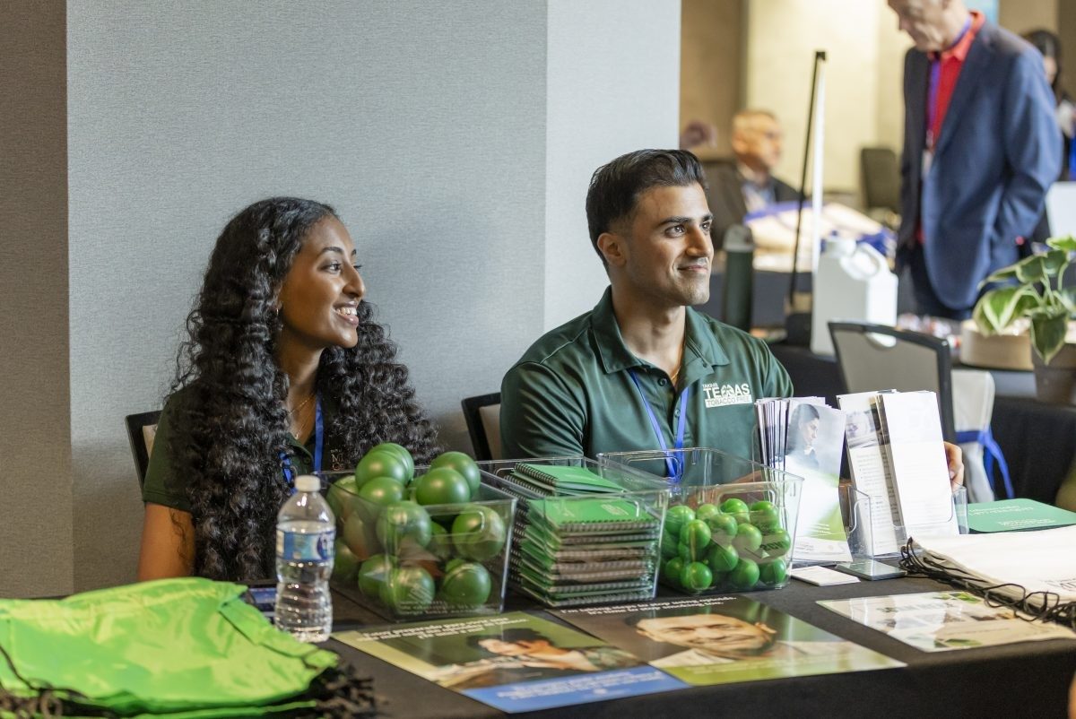 Two people seated behind a table, smiling and looking off to the right. The table holds swag, including green bags, stress balls, and notebooks.