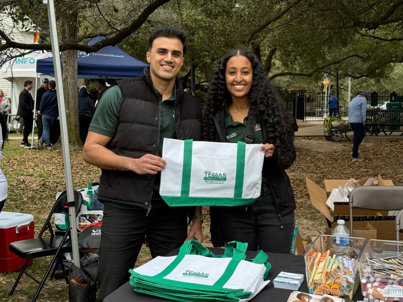 Two people at an outdoor conference table set-up holding a white bag with green straps that reads "Taking Texas Tobacco Free," with a pile of additional bags on the table