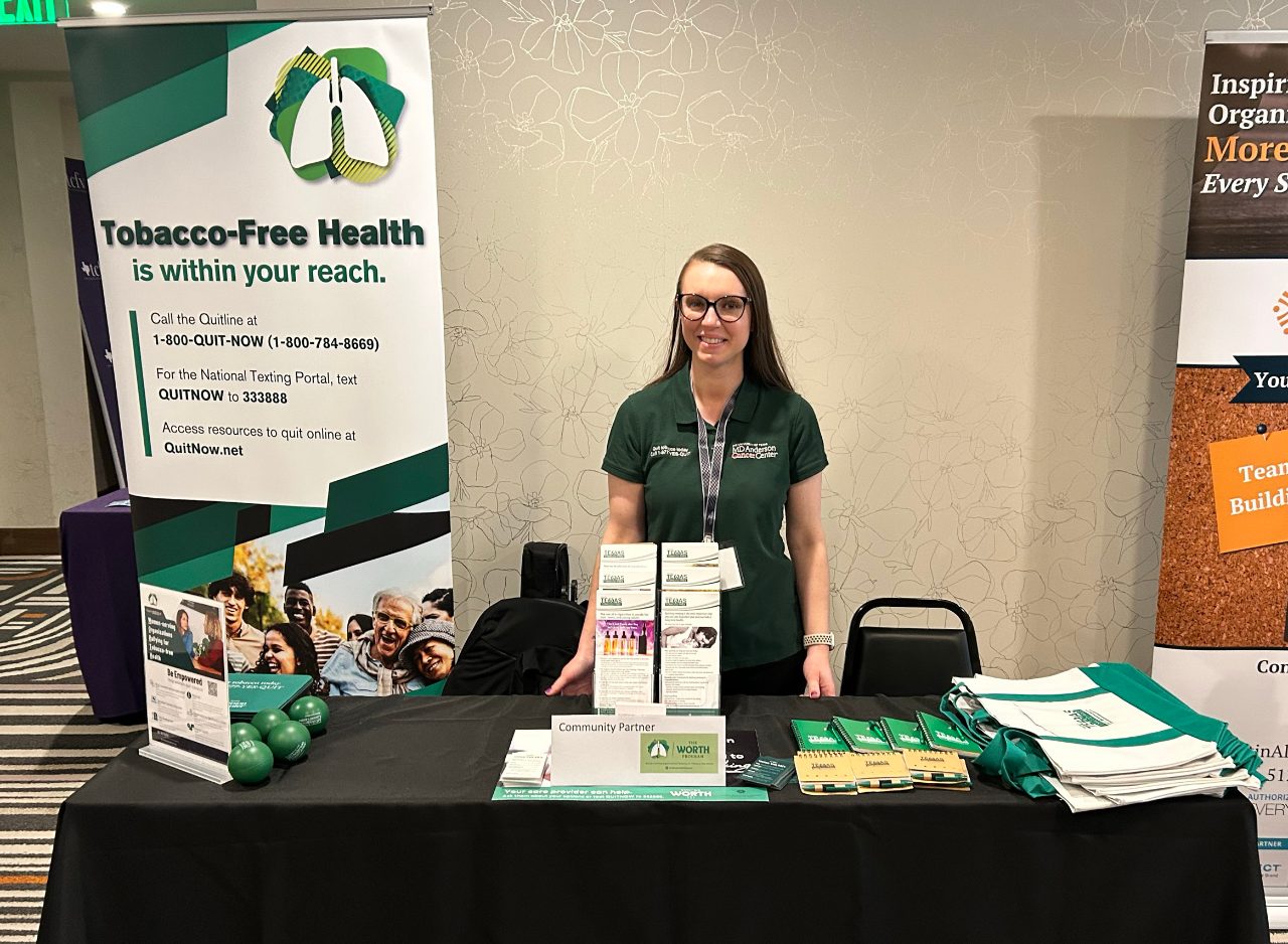 Woman stands behind a table in a conference exhibit hall with various swag on the table and a sign to her right that reads "Tobacco-Free Health is within your reach"