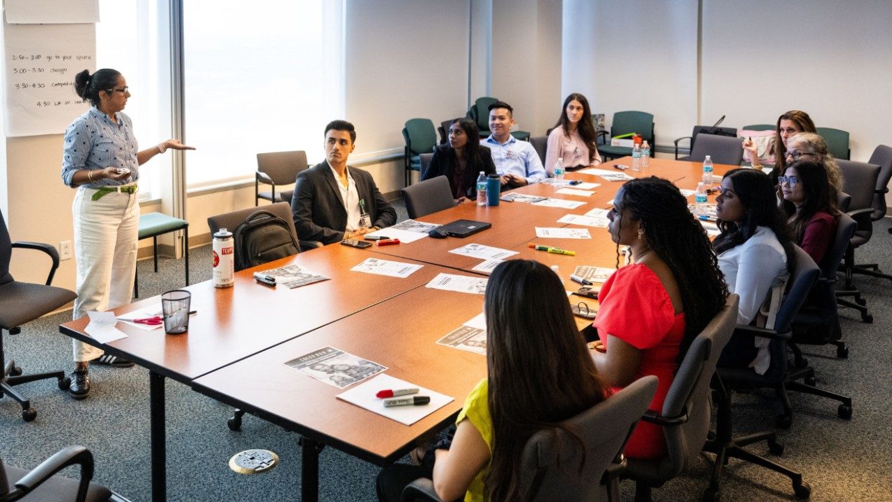 A group of people sit around a table and one person is standing and appears to be presenting