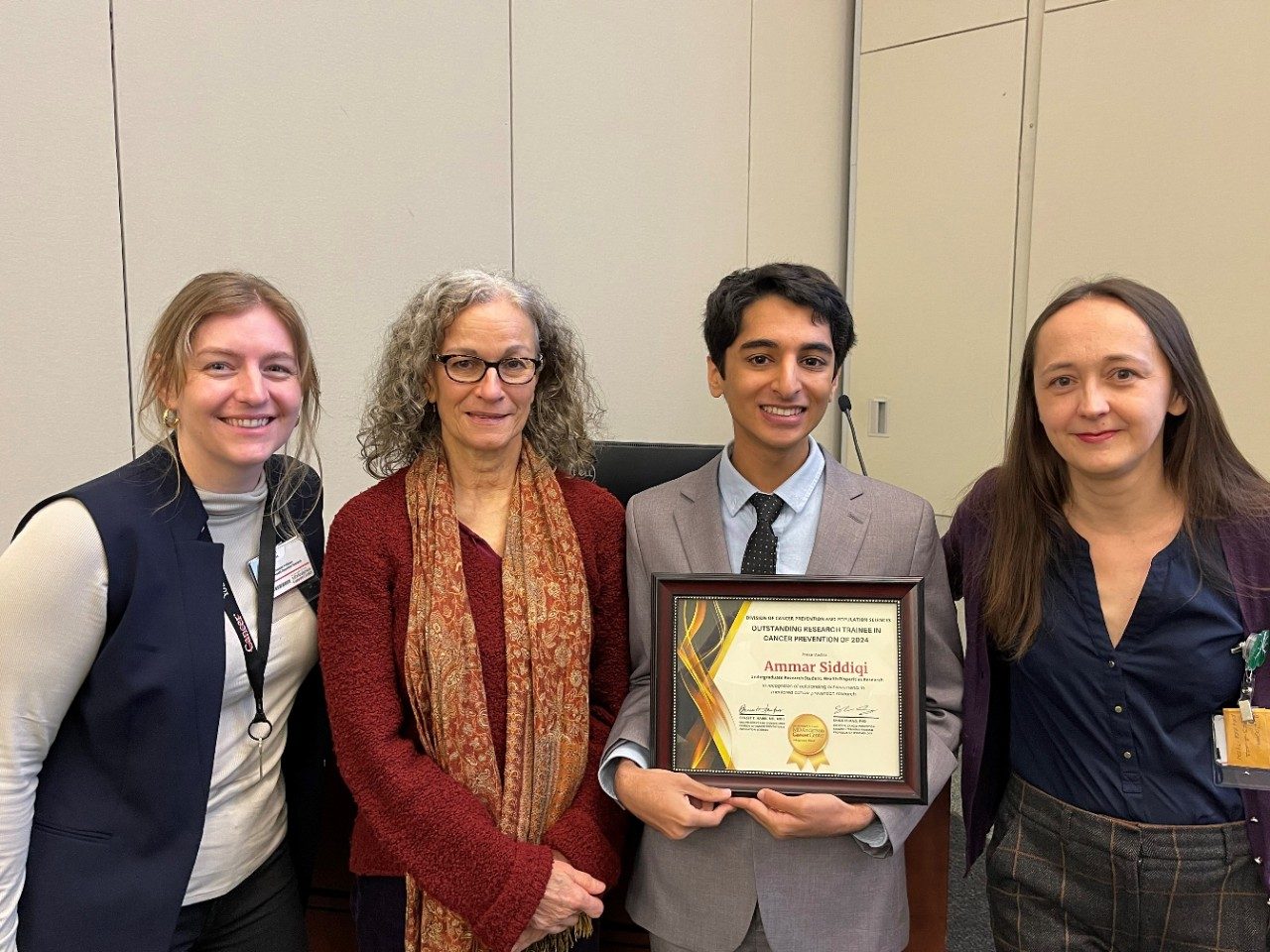 Young man in grey suit holding Outstanding Research Trainee in Cancer Prevention 2024 certificate with three women.