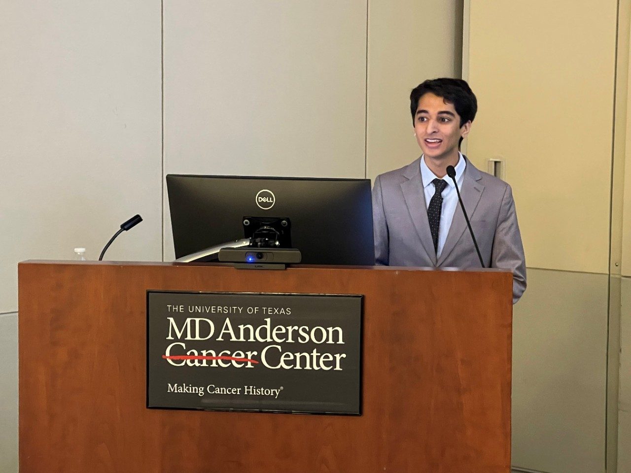 Young man in grey suit standing behind podium with MD Anderson Cancer Center sign, giving a talk.