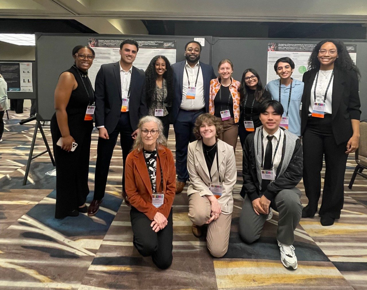11 people posed in front of a poster, with 8 in the back row and 3 kneeling on the floor in front