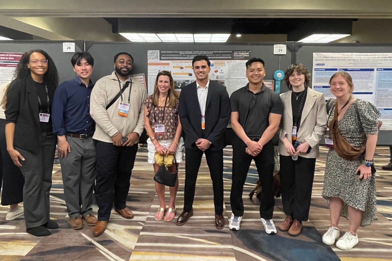 Eight people posed in a line in front of a poster at a conference