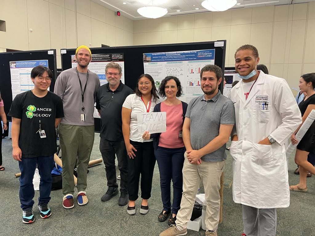 Seven people lined up in front of a poster, with one holding up a paper certificate