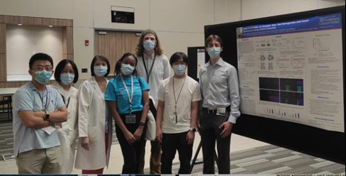 Seven lab members lined up with blue surgical masks next to a poster