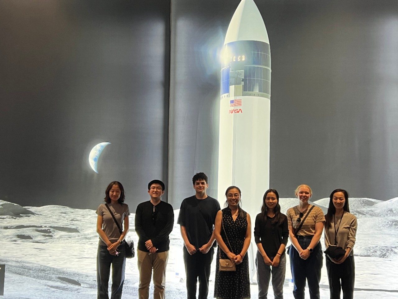 7 members of the Zhao Lab posing in front of a rocket on the moon