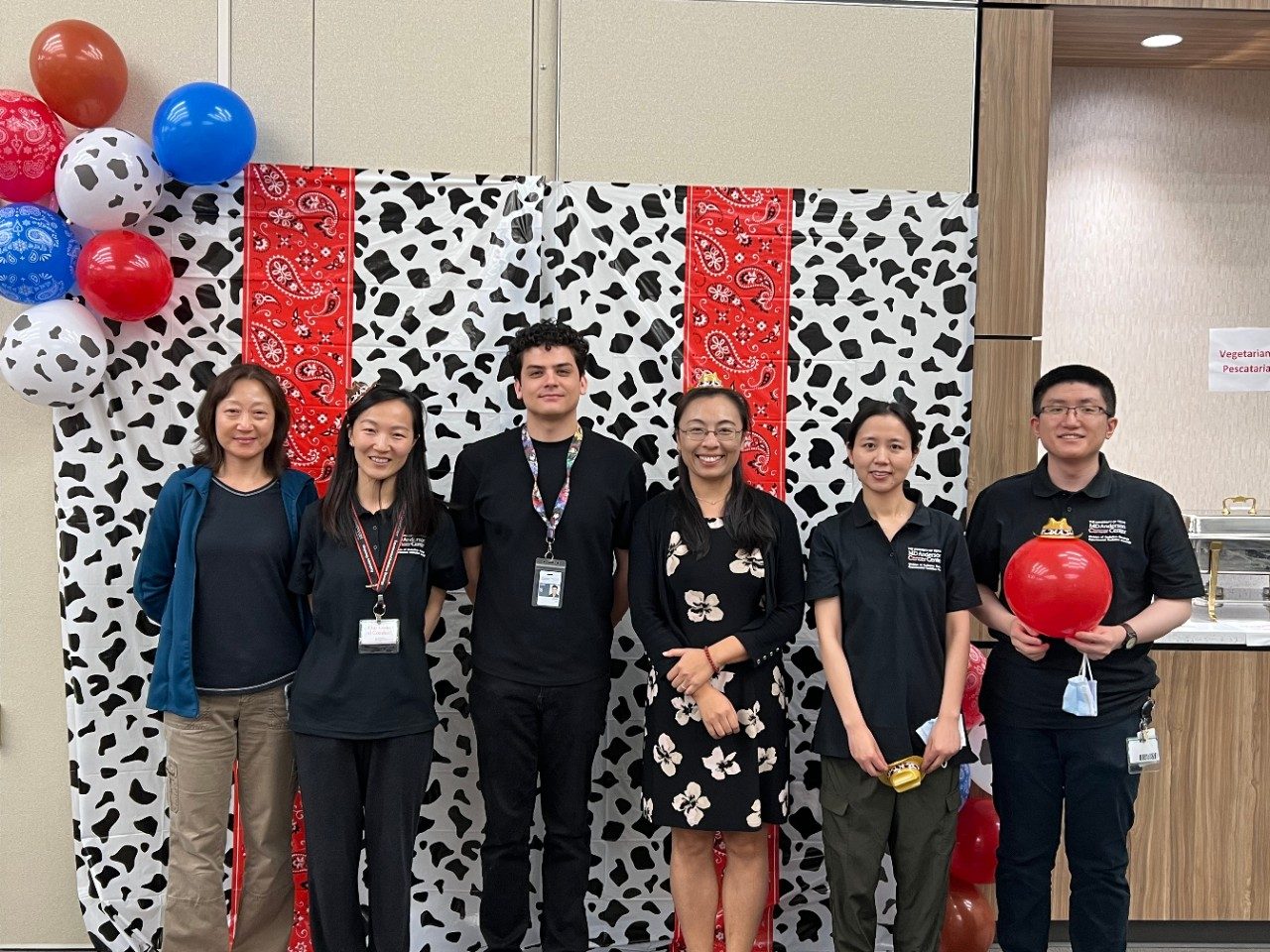 6 members of the Zhao Lab pose with miniature cowboy hats in front of a photo backdrop that is vertically striped with cow print and red paisley