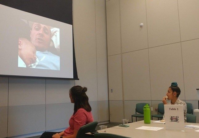 Two people watch a man on a screen with a baby