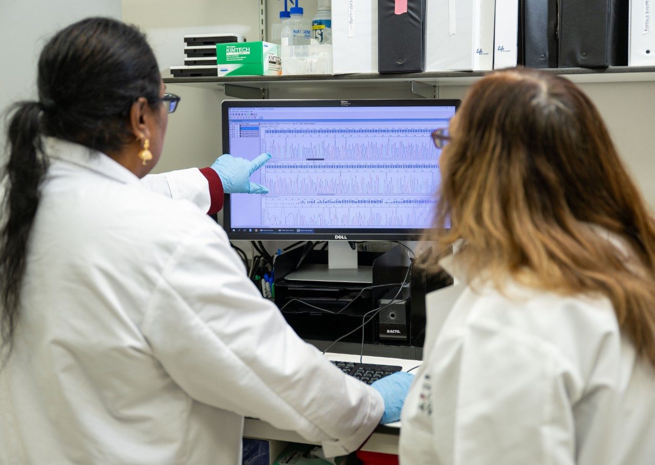Two people in white lab coats are standing at a computer workstation reviewing sequencing data displayed on a monitor. The screen shows multiple rows of peaks and waveforms, likely representing genetic sequences. Shelves with binders and lab supplies are visible in the background.