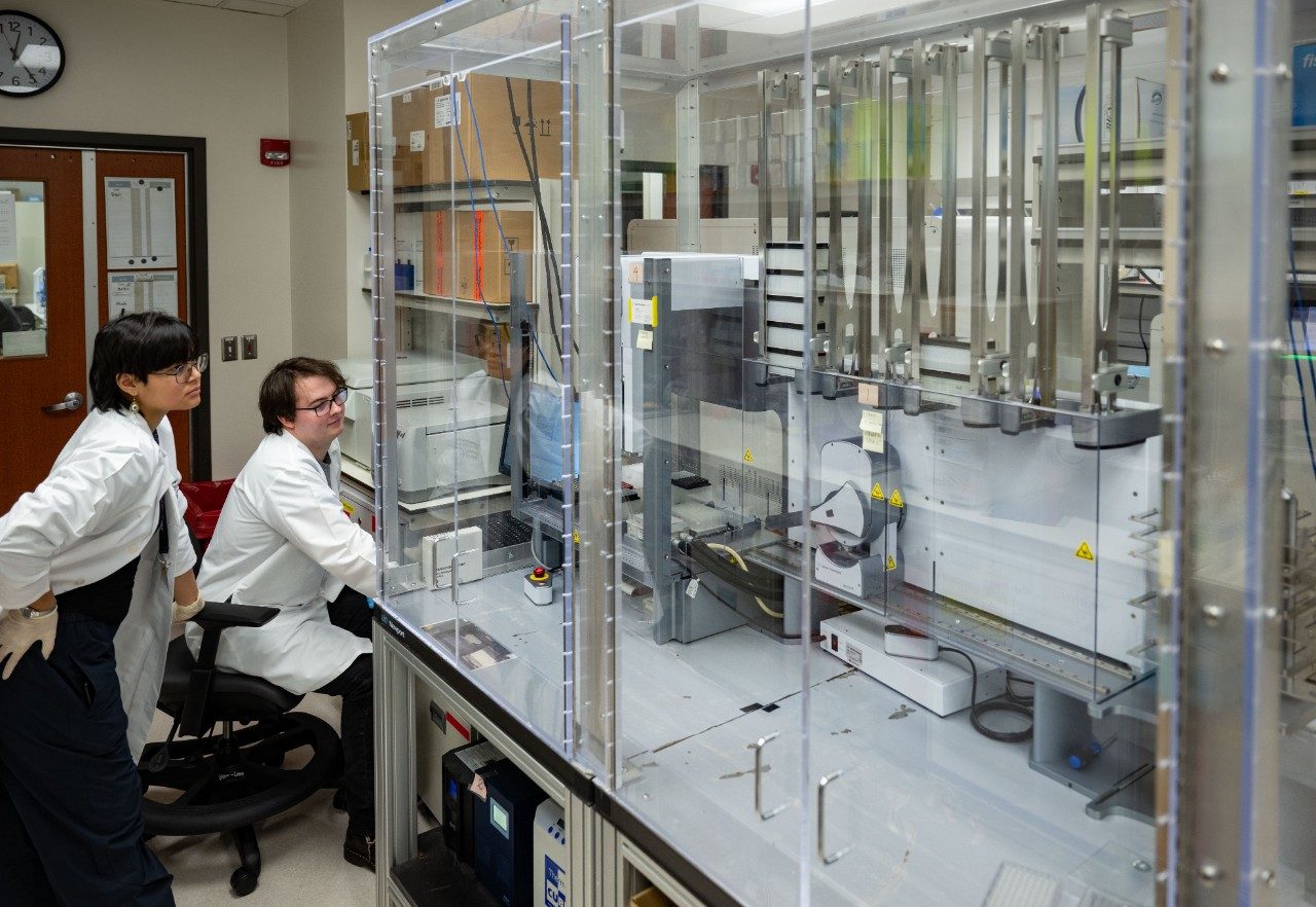 Two people in white lab coats are seated at a workstation in front of a large automated liquid handling system enclosed in glass. The machine contains multiple robotic arms and pipetting components. The background includes shelves with lab supplies and a door with posted notices.
