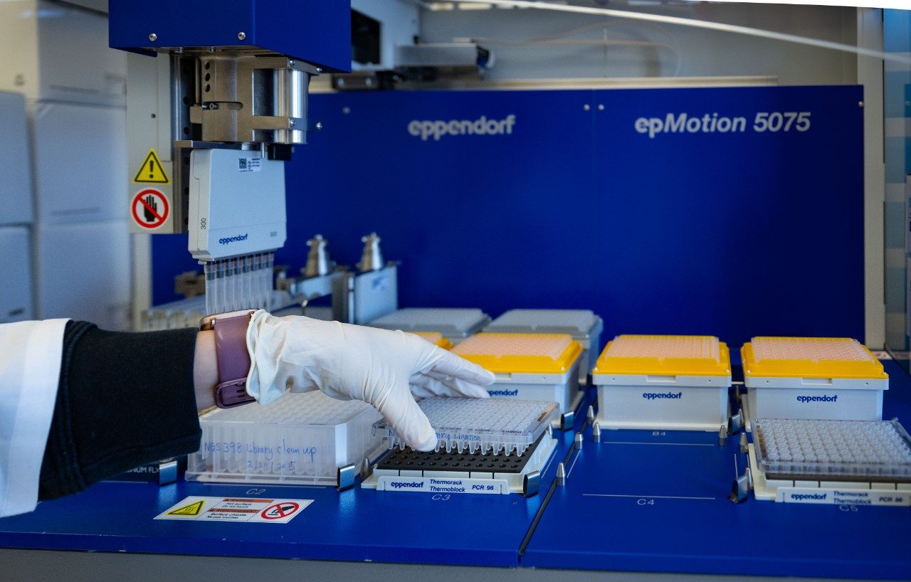Close-up of a gloved hand placing a sample plate into an automated liquid handling system labeled “Eppendorf epMotion 5075.” The machine has multiple racks with pipette tips and sample trays, and the background shows a blue enclosure with warning labels.