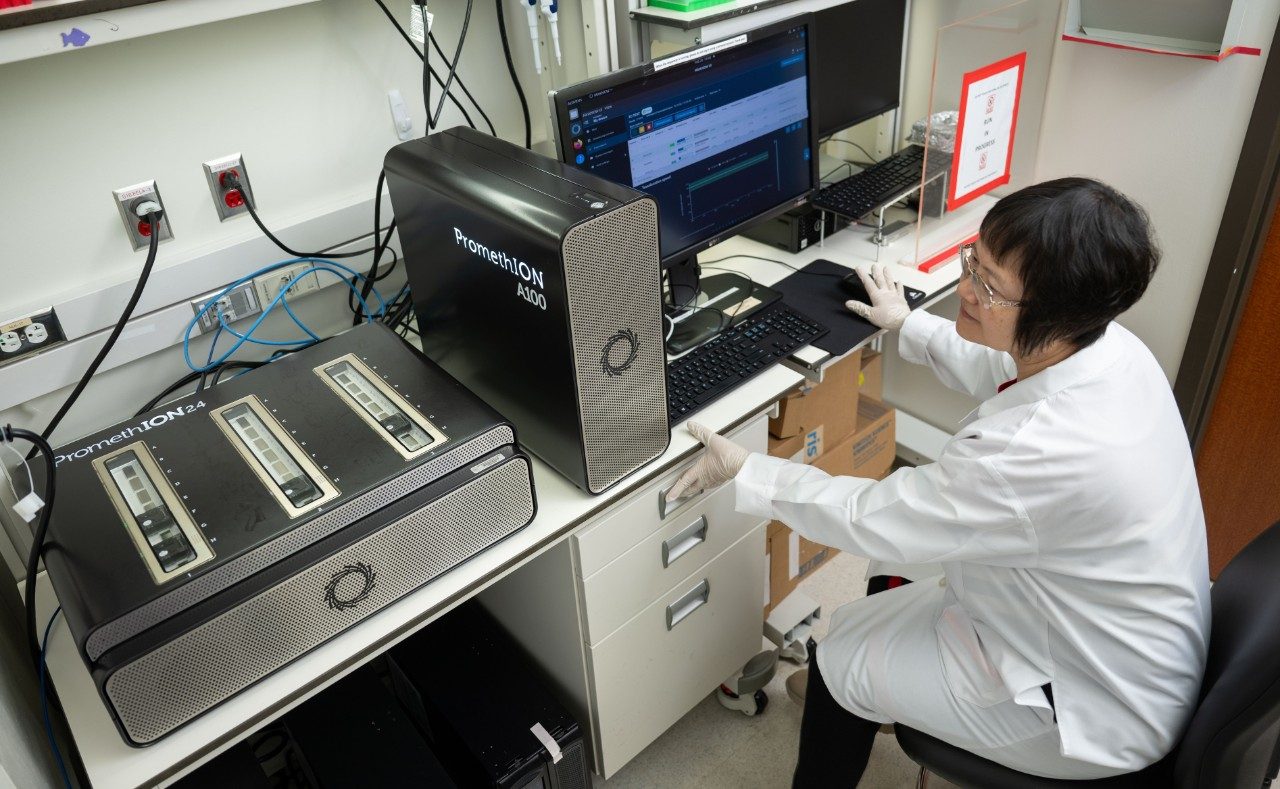 A person in a white lab coat is seated at a workstation operating a computer connected to two PromethION sequencing devices. The devices are black with metallic mesh panels and labeled “PromethION 24” and “PromethION 48.” The computer monitor displays sequencing software, and the desk has a keyboard, mouse, and cables.