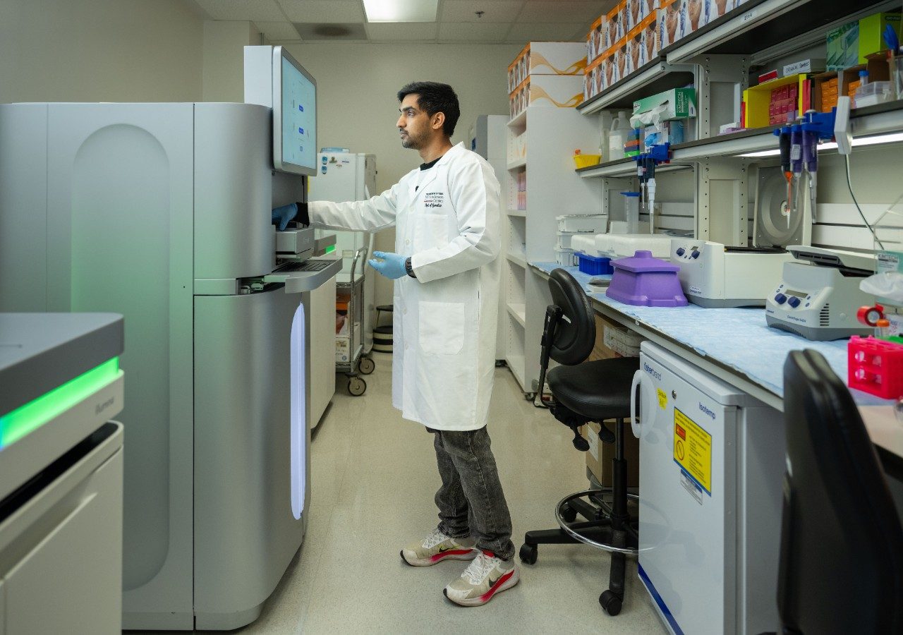 A person in a white lab coat and gloves is operating a large sequencing instrument in a laboratory. The machine has a vertical touchscreen interface, and the surrounding bench includes pipettes, racks, and other lab equipment.