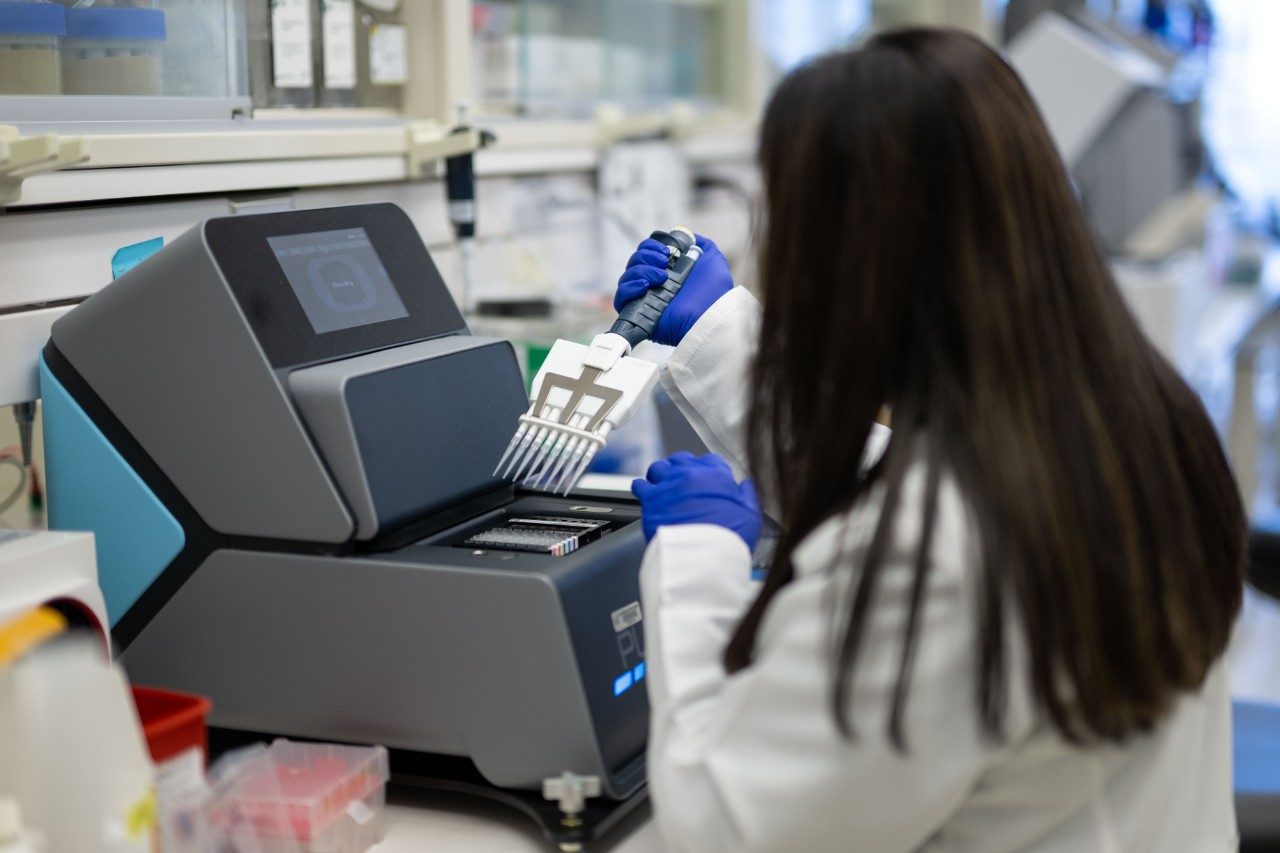 A person in a white lab coat and blue gloves is using a multichannel pipette to dispense liquid into a plate positioned inside a benchtop instrument. The machine has a touchscreen interface, and the surrounding bench includes racks and lab supplies.