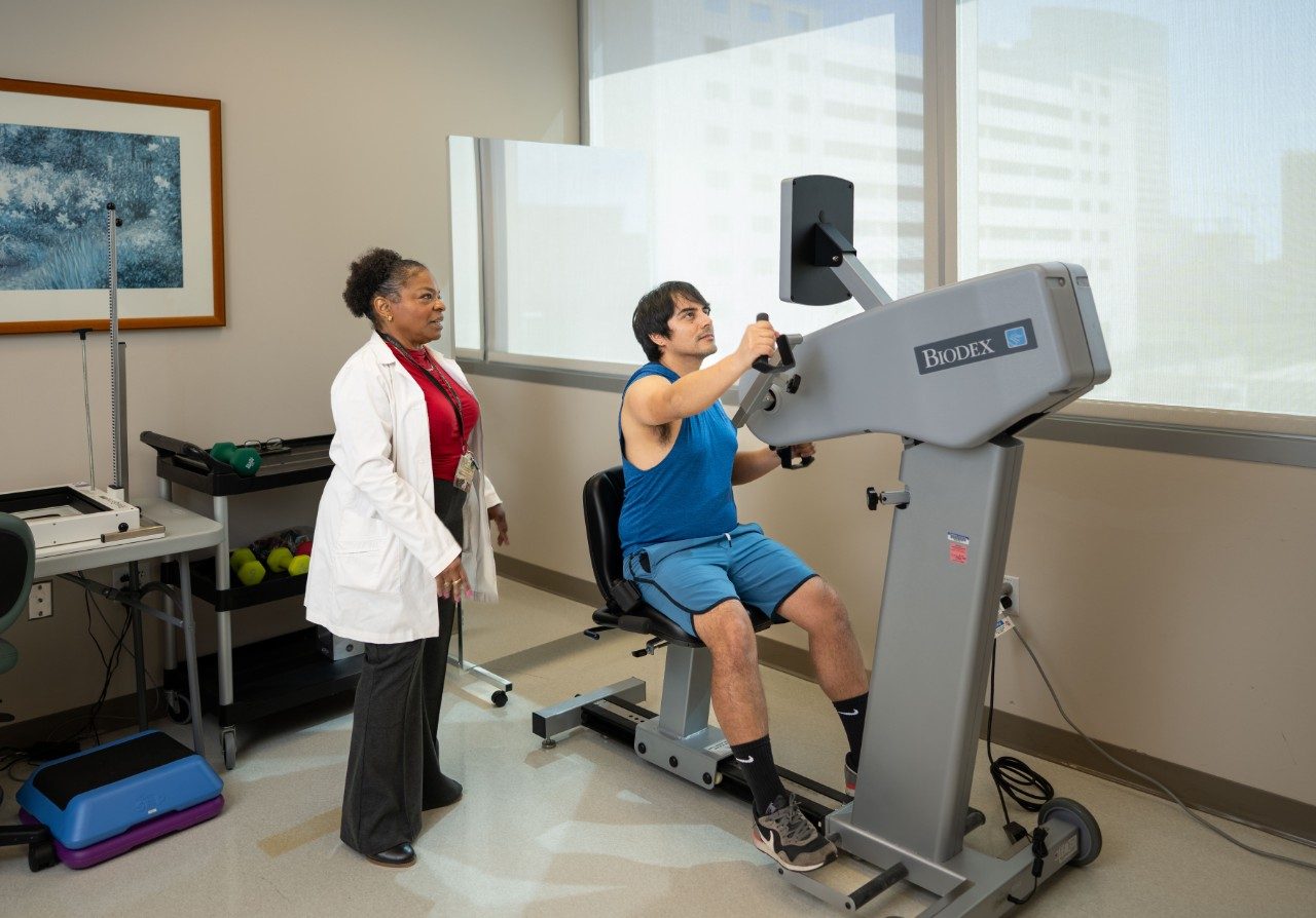 A person seated on a Biodex exercise machine performing an upper-body workout using rotating handles. Another person in a lab coat stands nearby supervising. The room has large windows and exercise equipment in the background.