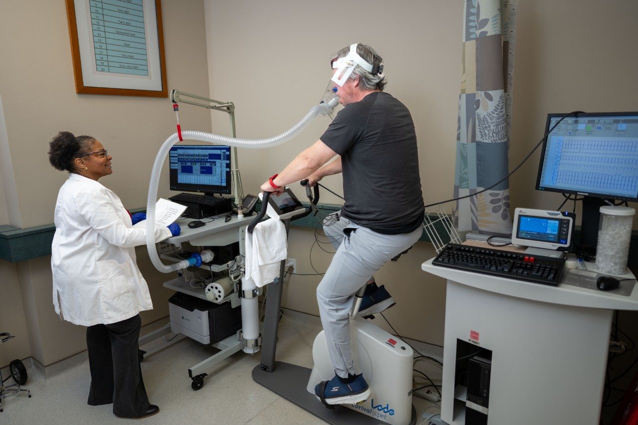 A person riding a stationary exercise bike while wearing a headpiece connected to breathing tubes for a fitness test. Another person in a lab coat stands nearby holding a sheet of paper. Computer monitors display physiological data.