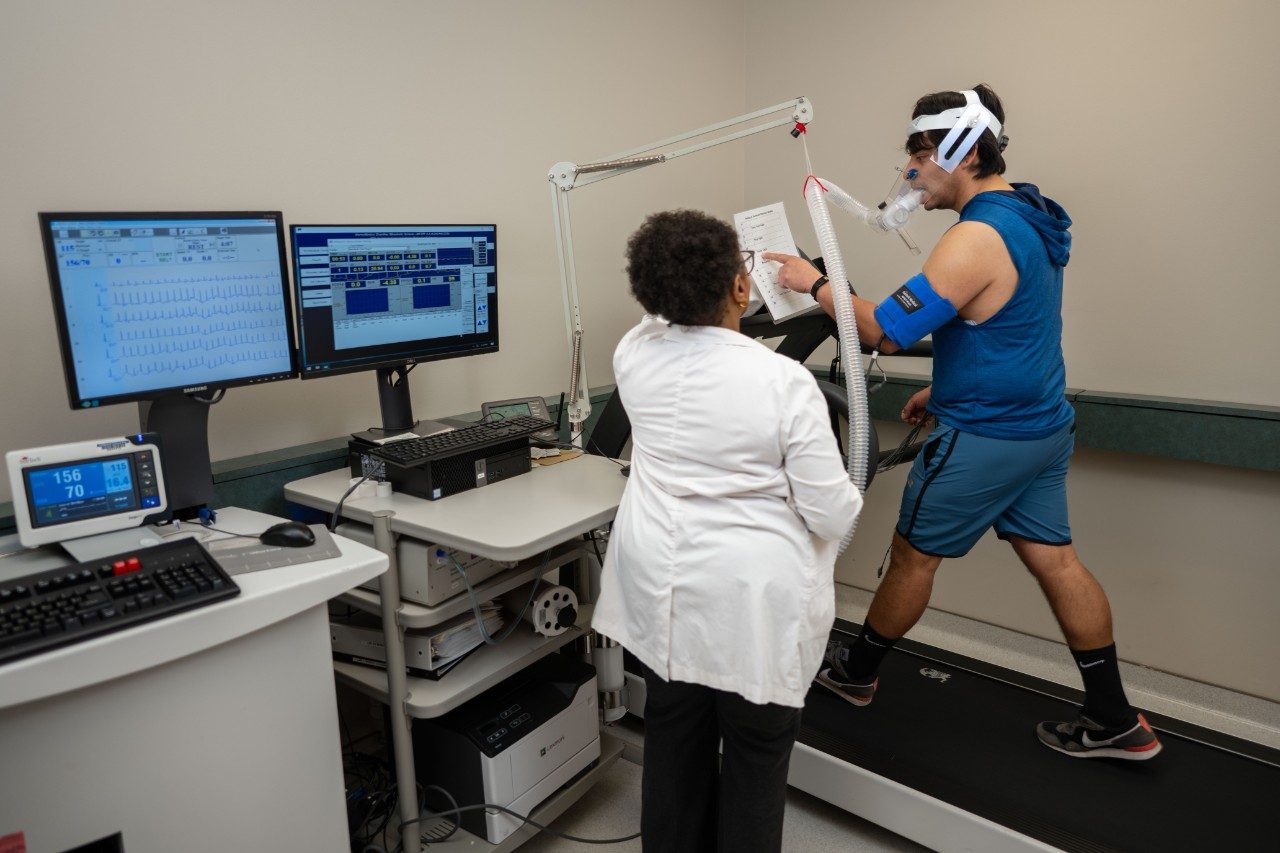 A person wearing athletic clothing and a headpiece with breathing tubes is walking on a treadmill during a fitness test. Another person in a white lab coat stands nearby monitoring the test. Two computer monitors display data, including heart rate and other measurements.
