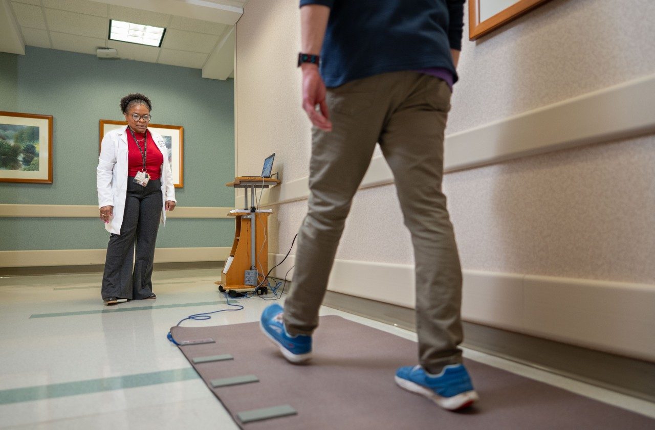 A person walking along a mat with sensors on the floor in a hallway, while another person in a lab coat stands at a distance near a small computer station. Framed artwork hangs on the walls.