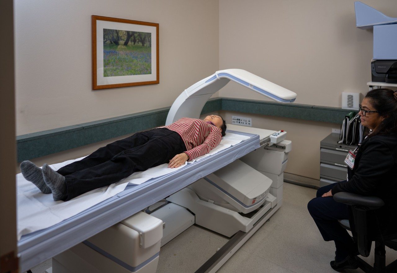 A person lying flat on a padded table under a large medical imaging scanner arm in a clinical room. Another person sits nearby observing the procedure. Cabinets and a framed landscape picture are visible in the background.
