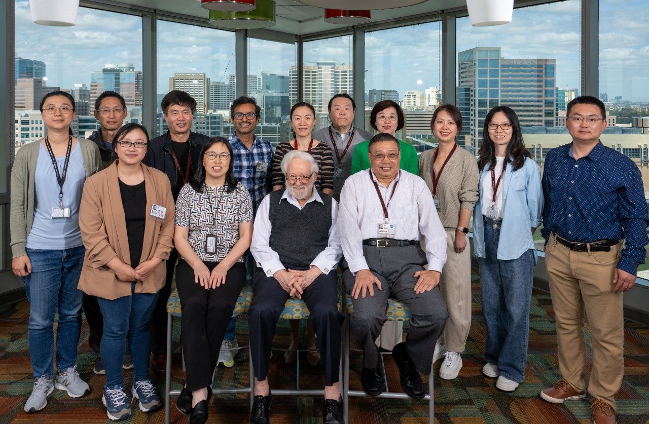 BISR group staff photo. A group photo of thirteen people standing and sitting in a room with large windows overlooking a city skyline. The group is arranged in two rows, with two people seated in front and the rest standing behind. The floor has a patterned carpet, and ceiling lights are visible.