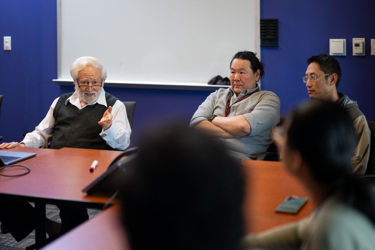 Five people seated side by side at a conference table in a meeting room. The background includes a white board and a blue wall. The table is empty except for a few small items.