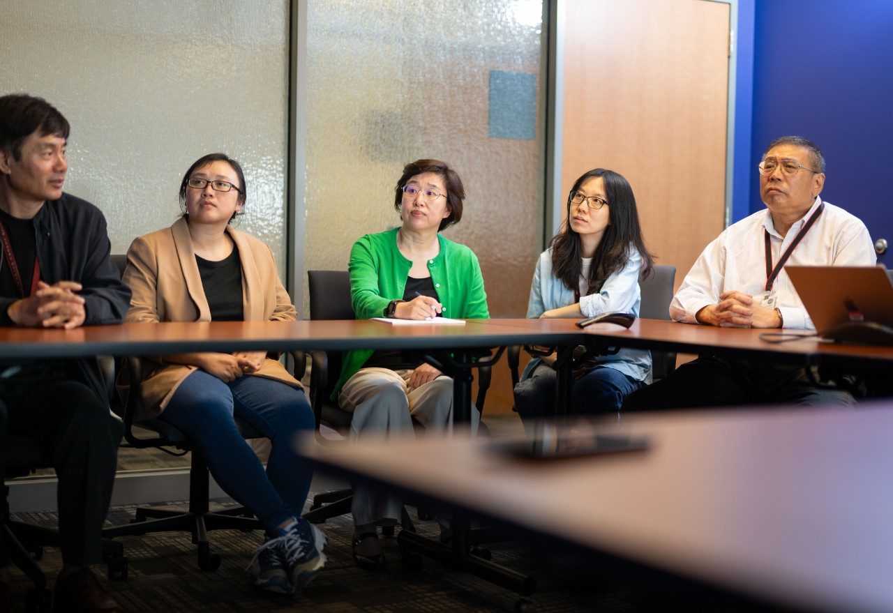 Five people seated side by side at a conference table in a meeting room. The background includes frosted glass panels and a blue wall. The table is empty except for a few small items.