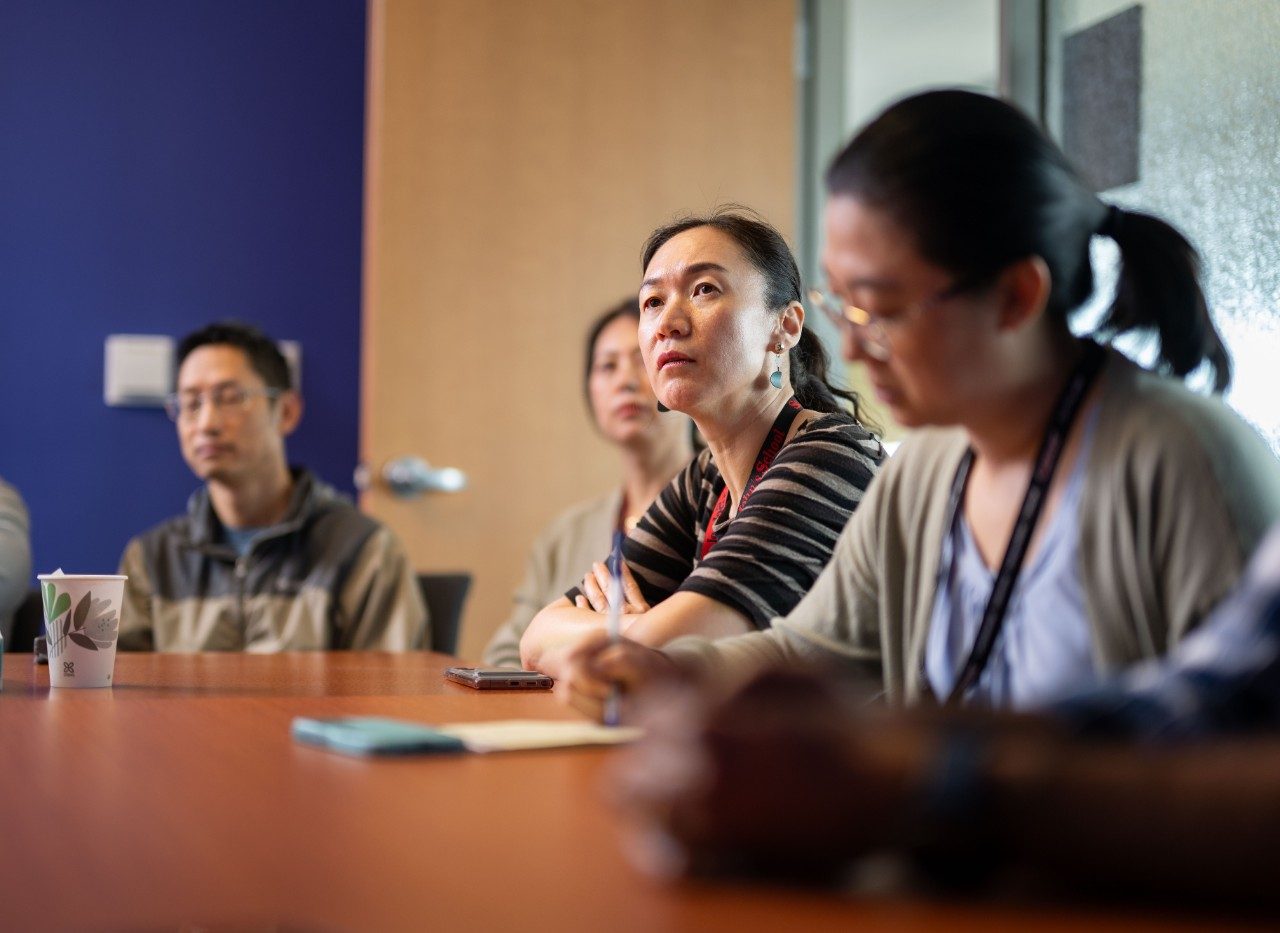 Close-up view of several people seated at a conference table during a meeting. A notepad and pen are visible on the table, along with a paper cup. The background includes a blue wall and a wooden door.