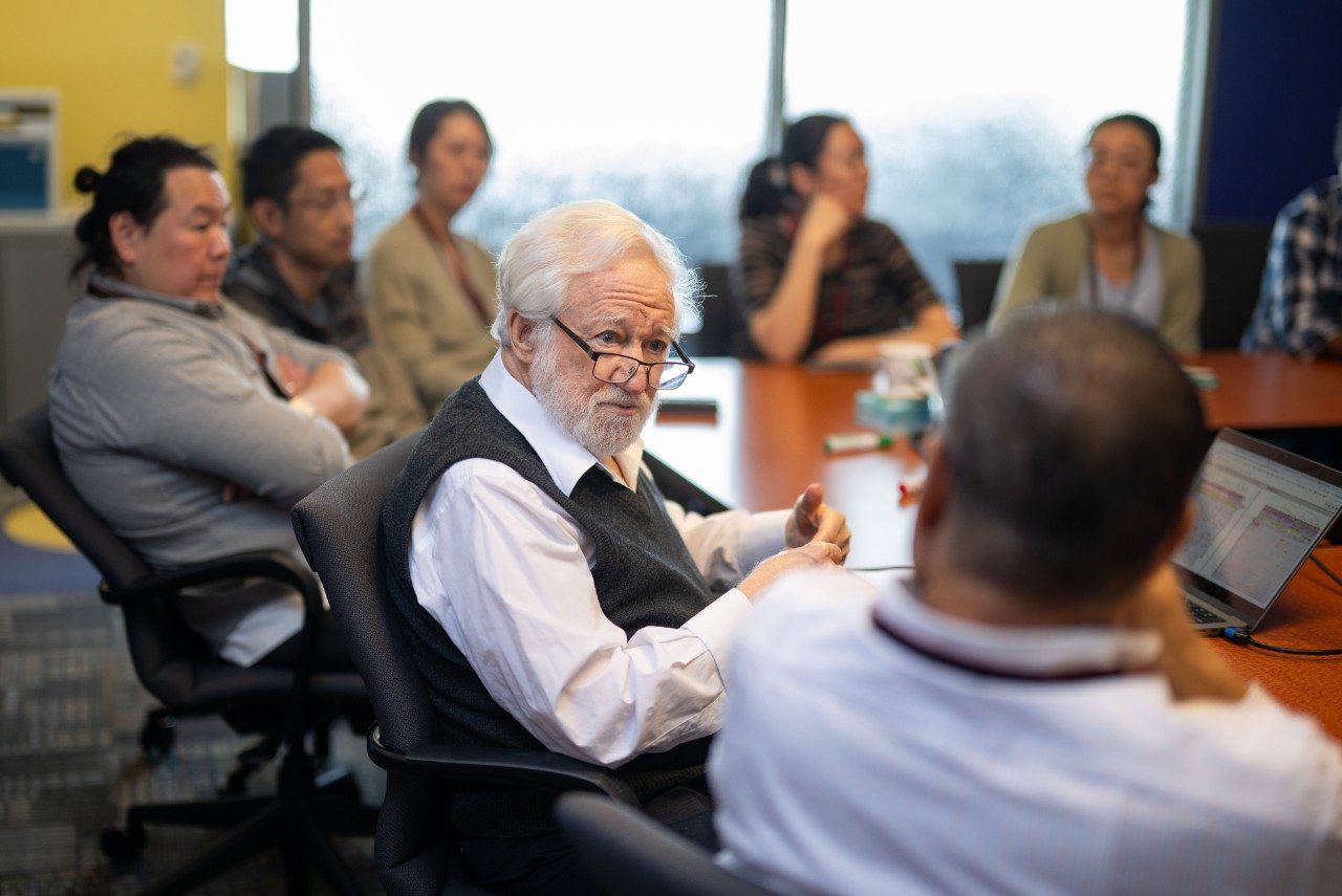 A group of people seated around a large conference table in a meeting room. One person in the foreground is speaking while others listen. A laptop is open on the table, and a large window in the background lets in natural light.