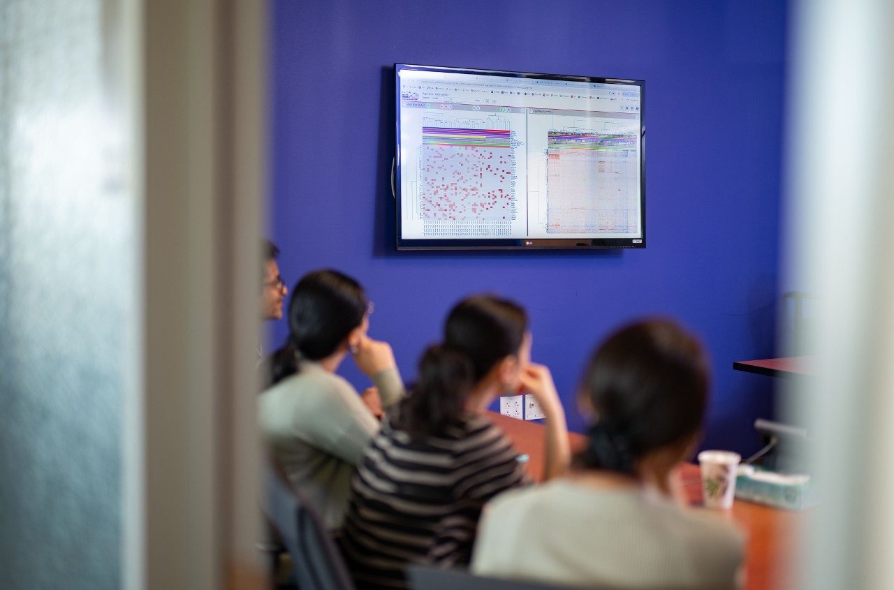 People seated at a conference table looking at a wall-mounted screen displaying charts and data visualizations. A paper cup and other small items are on the table.