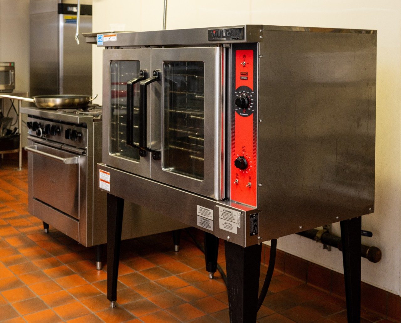 A commercial stove with a frying pan on the burner. A large stainless steel oven is positioned next to the stove, and the kitchen floor is covered with red tiles.