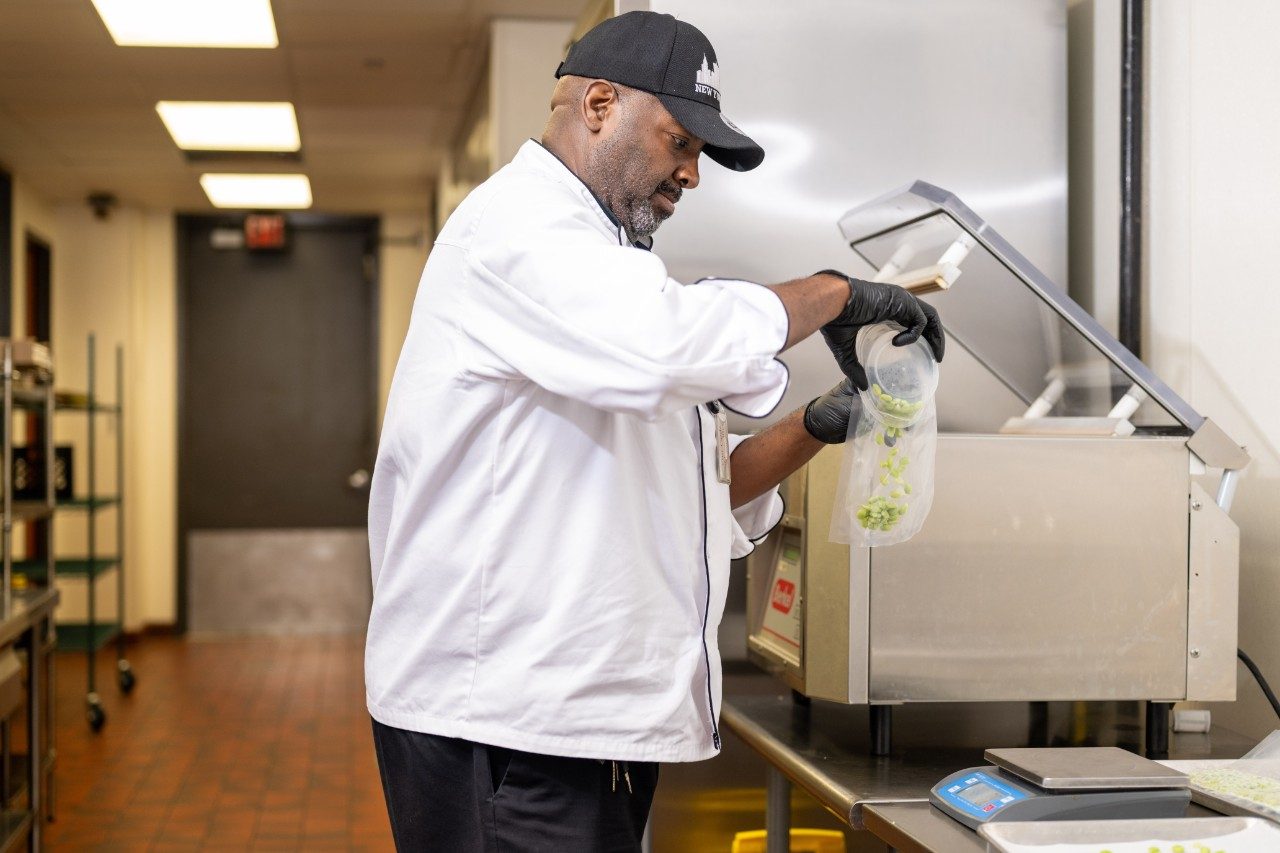 A chef in a white uniform and black gloves is pouring edamame from a plastic bag into a large stainless steel machine in a commercial kitchen. A digital scale is visible on the counter nearby.