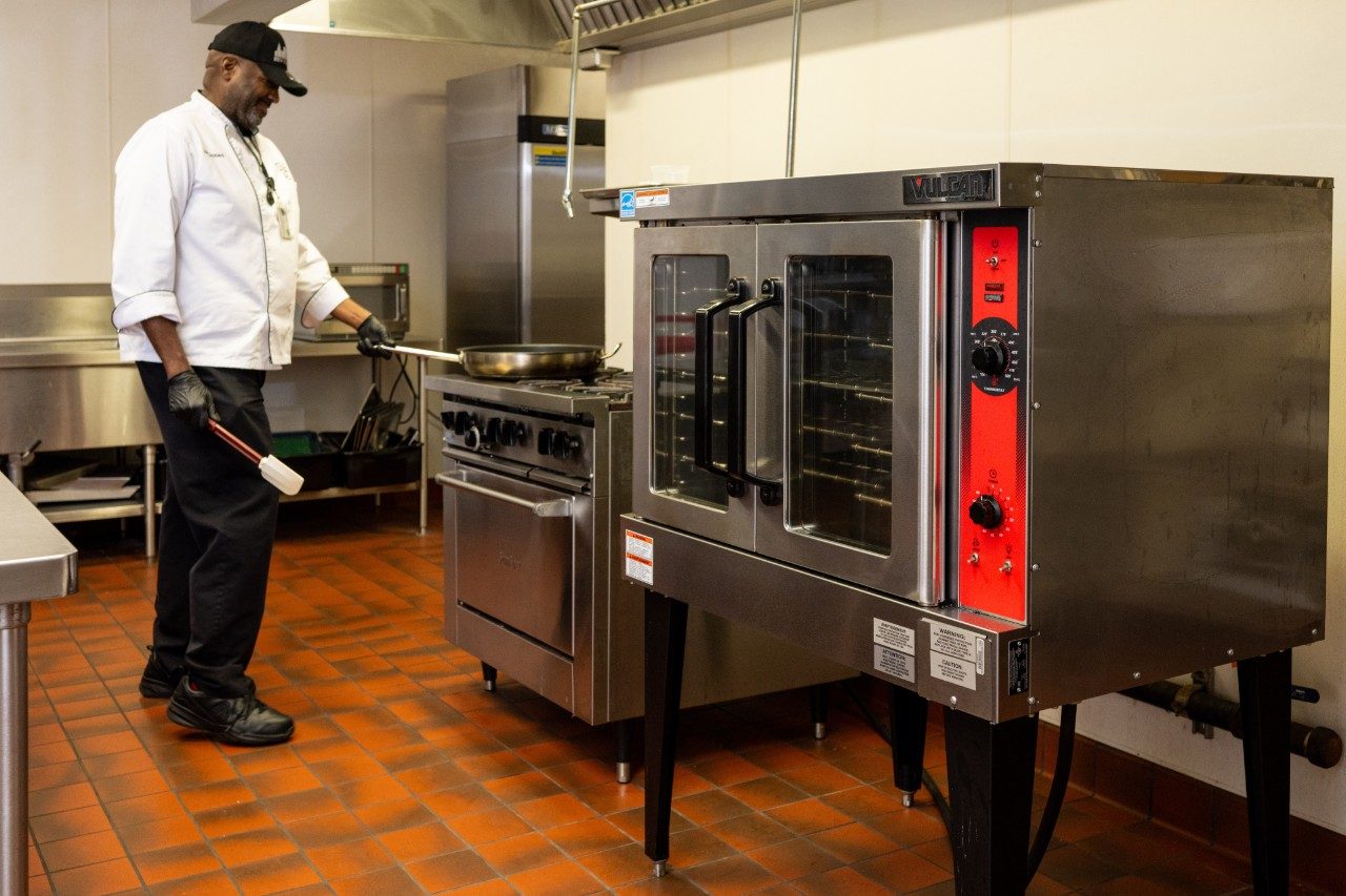 A chef in a white uniform and black gloves is standing in front of a commercial stove with a frying pan on the burner. A large stainless steel oven is positioned next to the stove, and the kitchen floor is covered with red tiles.
