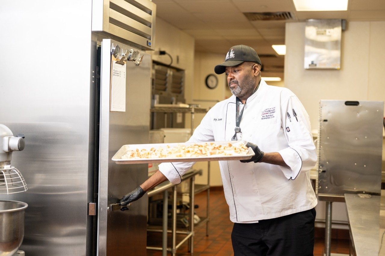 A chef wearing a white uniform and black gloves is holding a large tray of prepared food while opening a stainless steel refrigerator door in a commercial kitchen. The background includes metal shelving, a stand mixer, and other kitchen equipment.