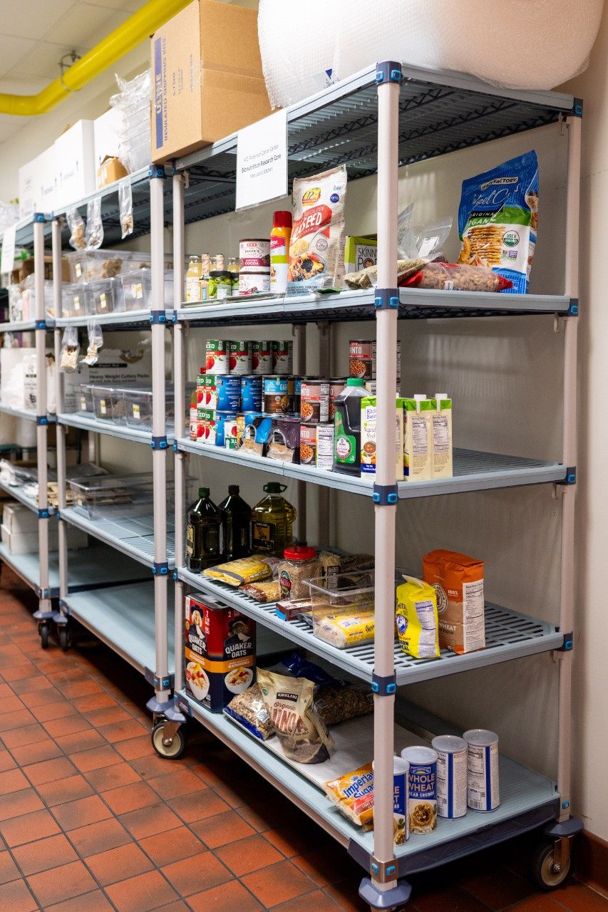 Two metal shelving units in a kitchen storage area stocked with various food items and supplies. The shelves hold canned goods, cooking oils, spices, pasta, and packaged snacks. Clear plastic bins and boxes are stored on the top shelves.