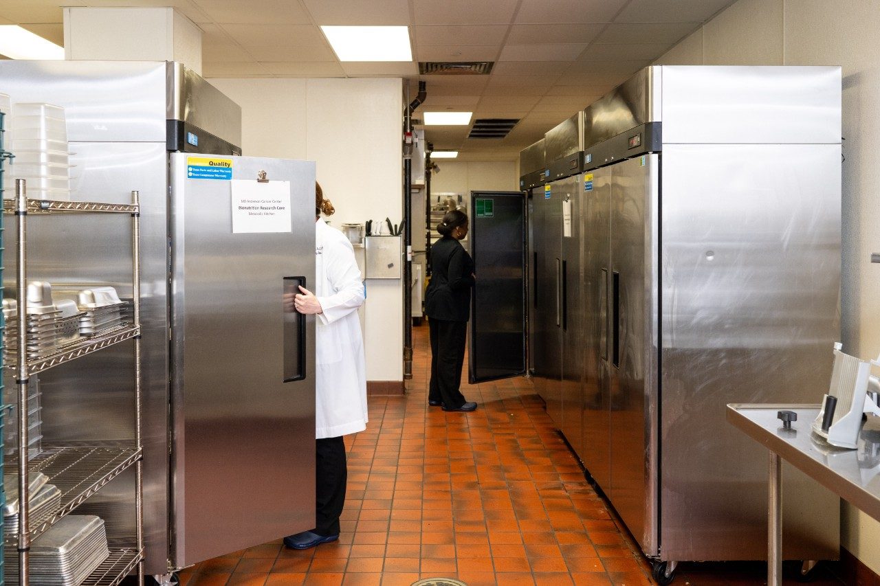A commercial kitchen hallway with multiple stainless steel refrigerators lined up along the wall. One person is opening a refrigerator door, and another person is standing near the far end of the hallway. Metal shelving with trays and dishes is visible on the left.