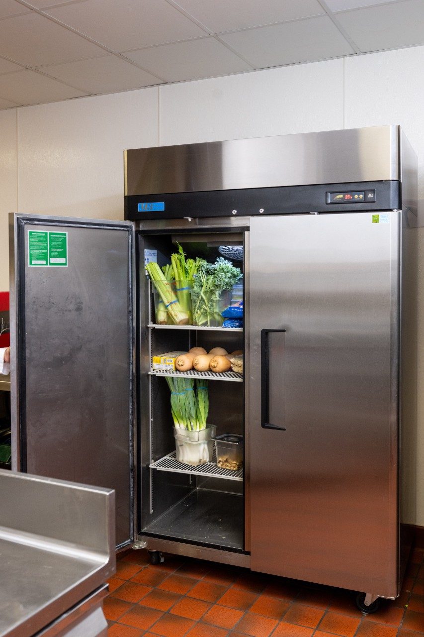 An open stainless steel refrigerator in a commercial kitchen showing shelves stocked with fresh vegetables, including celery, onions, and leafy greens. The refrigerator has a digital temperature display at the top.