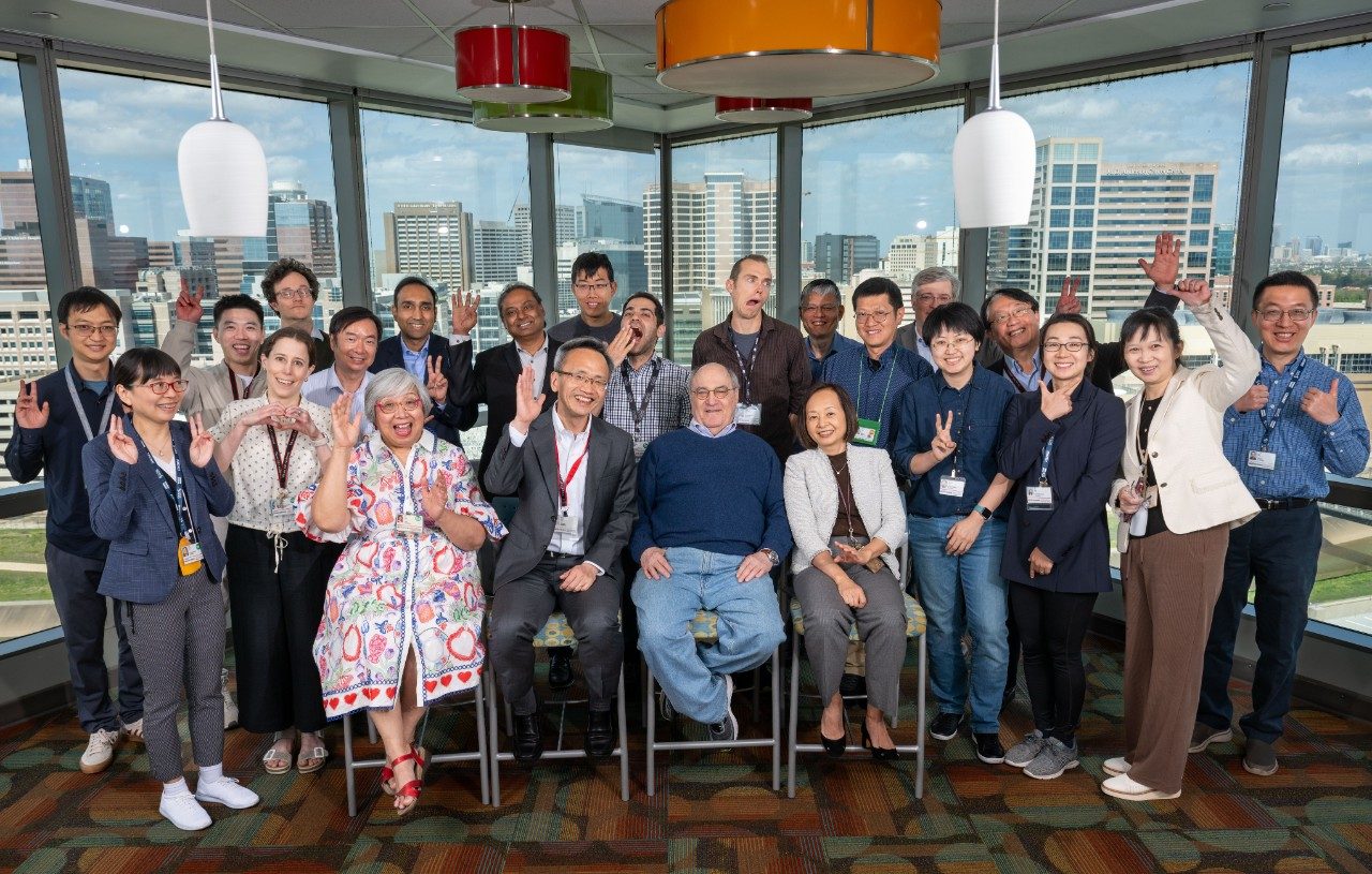 BRG group photo. Group photo of approximately twenty people standing and sitting in front of large windows with a city view. Several individuals are making peace signs with their hands. The room has colorful hanging light fixtures and patterned carpet.