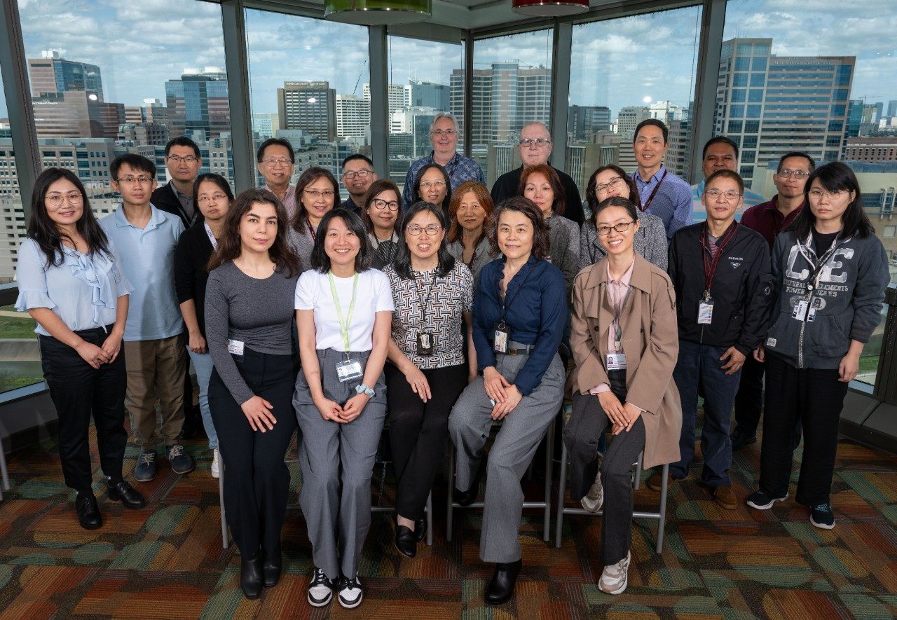 BRG group photo. Group photo of approximately twenty people standing and sitting in a room with large windows overlooking a city skyline. The group is arranged in two rows, with some seated on stools and others standing behind. The floor has a patterned carpet.