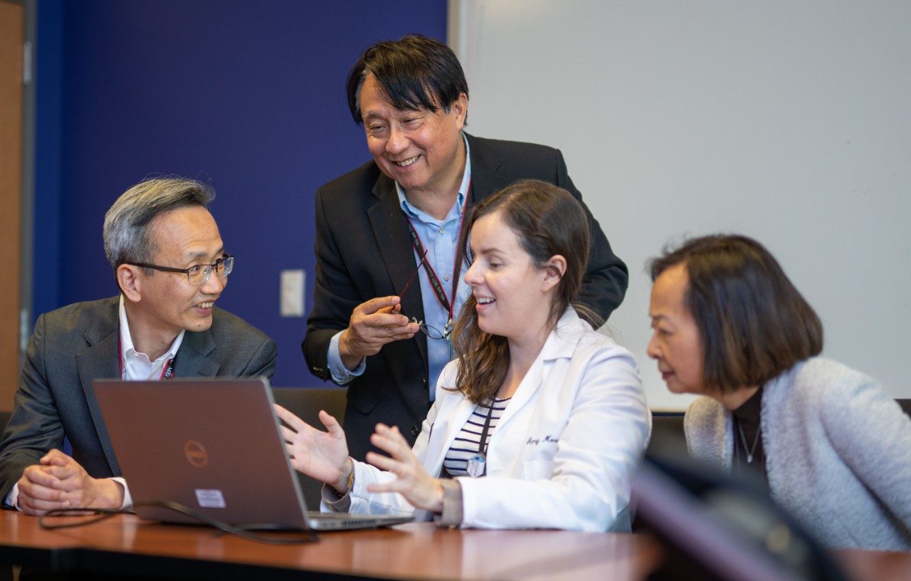 Four people seated and standing around a conference table in a meeting room. A laptop is open on the table, and one person is gesturing while speaking. The background includes a blue wall and a whiteboard.