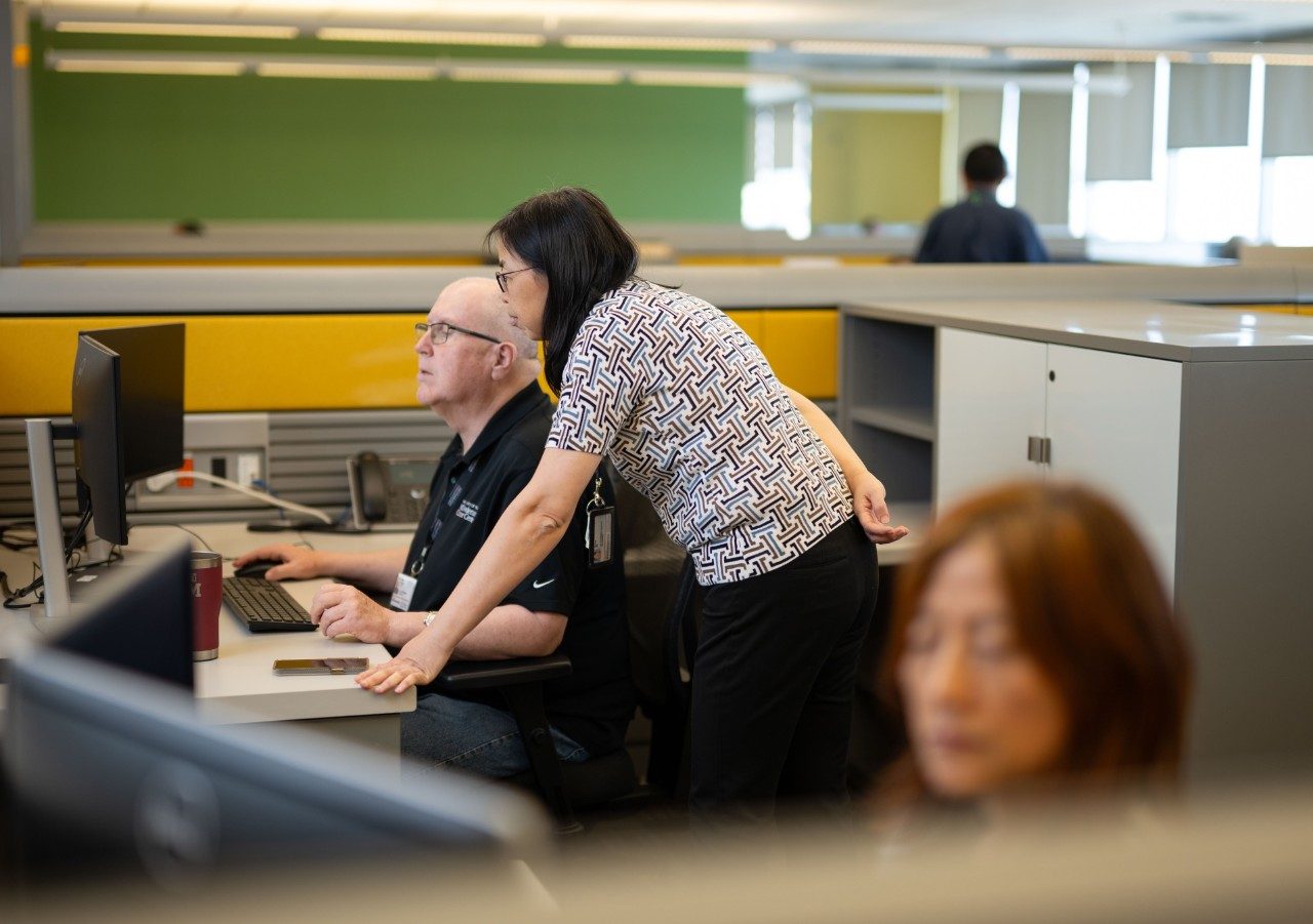 Two people in an open office space looking at a computer monitor. One person is seated at the desk while the other is leaning over, pointing at the screen. The workspace includes cubicles with yellow panels and multiple monitors. Another person at a desk in the foreground.