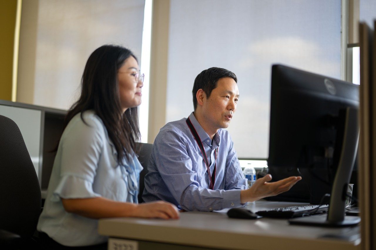 Two people seated at a desk in an office, looking at a large computer monitor. The background includes windows with blinds partially drawn, allowing natural light into the room.