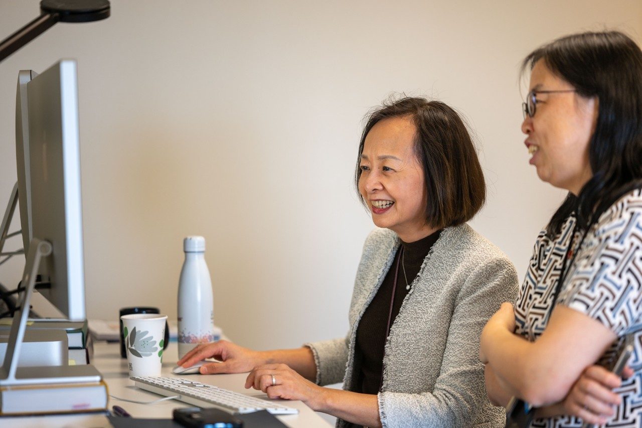 Two people working together at a desk with a computer monitor. One person is seated typing on a keyboard, while the other stands nearby observing. A coffee cup and water bottle are on the desk.