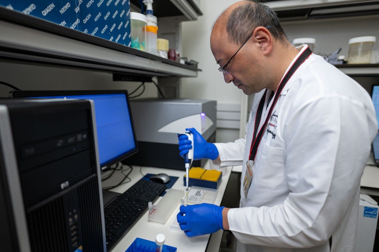 A person wearing a white lab coat and blue gloves is using a pipette to transfer liquid into a container in a laboratory setting. The workstation includes a computer monitor displaying data, a keyboard, and laboratory equipment such as racks with tubes and a benchtop instrument.