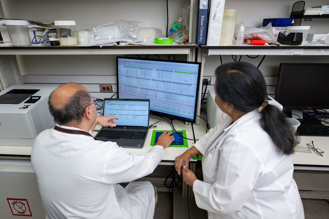 Two individuals in white lab coats are seated at a laboratory workstation, reviewing data on a laptop and a large monitor. The screen shows multiple rows of graphs or sequencing data. The bench is cluttered with lab supplies and equipment.