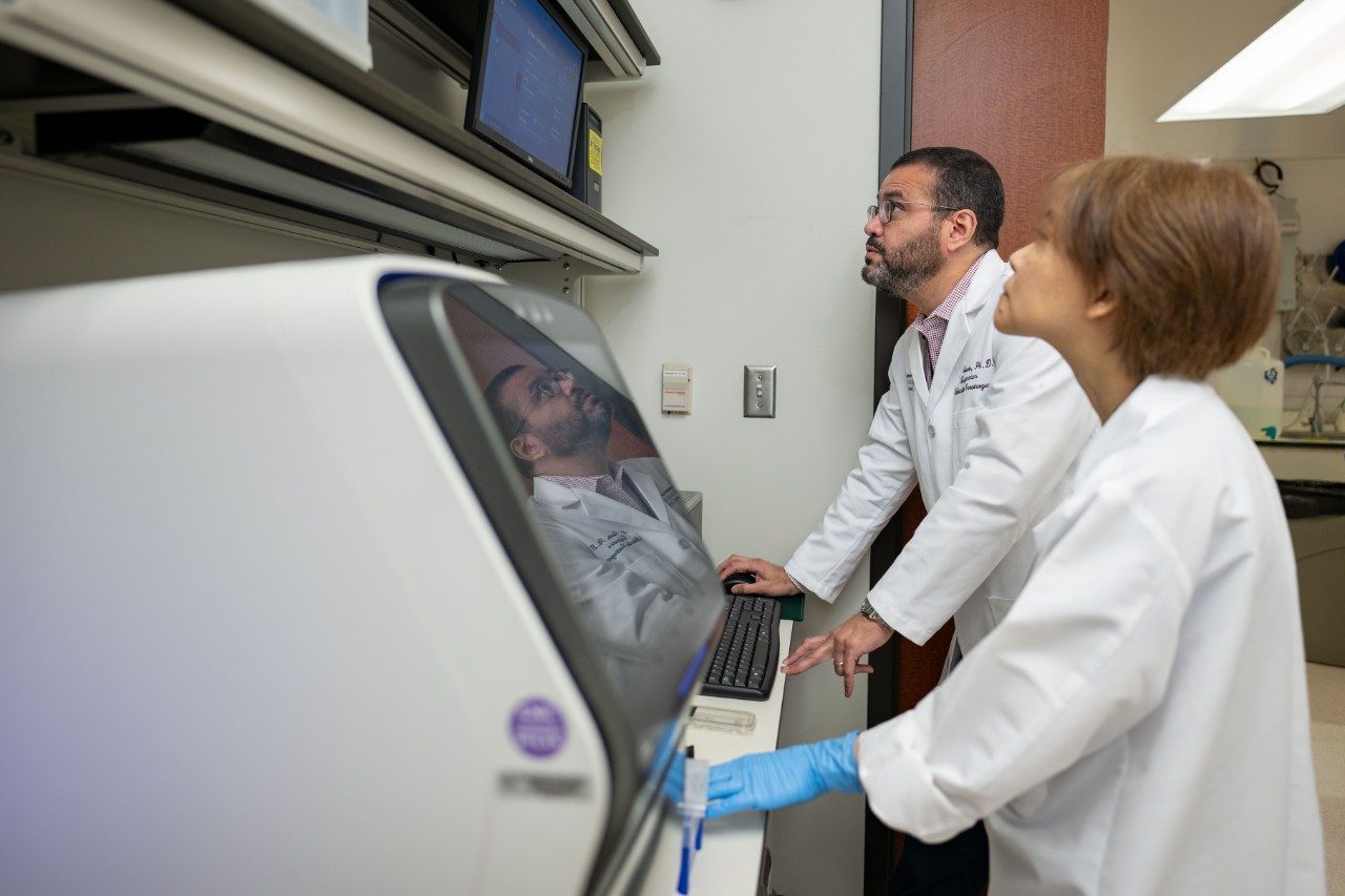 Two individuals in lab coats are working at a large sequencing instrument in a laboratory. One person is typing on a keyboard connected to the machine, while the other stands nearby observing.