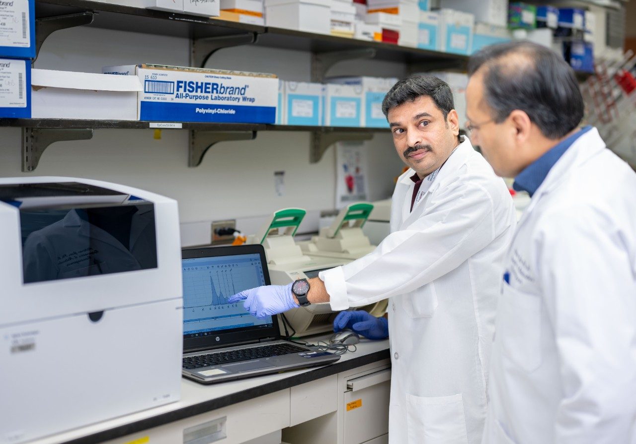 Two individuals in lab coats are reviewing data on a laptop in a laboratory. The laptop screen shows a graph or sequencing data, and lab equipment such as a benchtop analyzer and pipette tip boxes are visible in the background.
