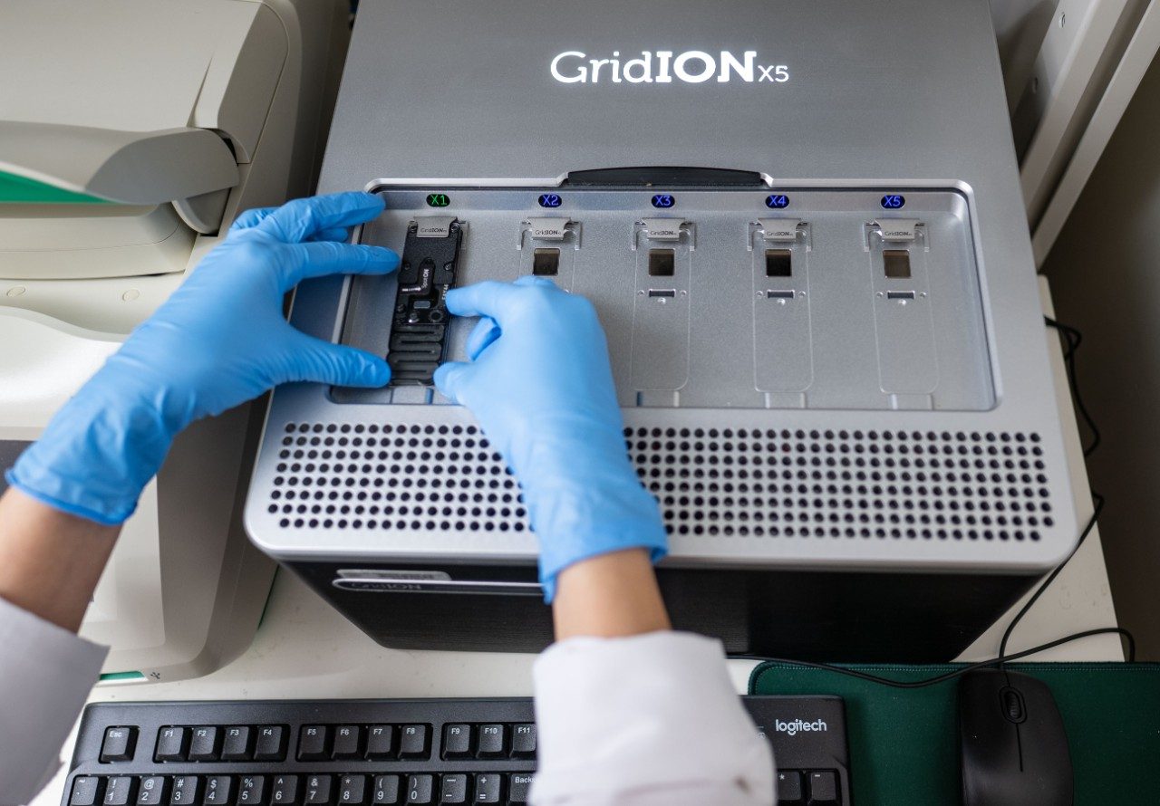 Close-up of gloved hands loading a sample cartridge into a GridION X5 sequencing device. The machine has five slots for cartridges and is positioned above a computer keyboard.