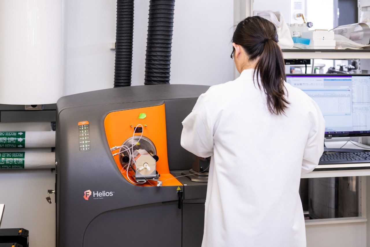 A person in a white lab coat is working with a Helios instrument, which has an orange panel and tubing visible. A computer monitor displaying data is positioned on the right side of the workstation.